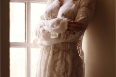 Sensual boudoir portrait of a sexy young woman in sheer white lace nightgown over naked body standing by the window